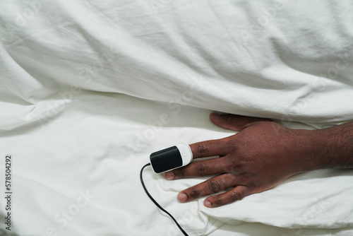 Hand of young African American male patient with pulse oximeter on fingertip lying under white cotton sheet in bed in hospital ward photo