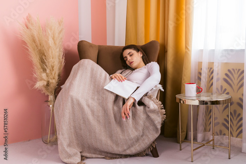 A portrait of a beautiful, long-haired girl who fell asleep in a chair after reading a book and covered herself with a blanket.