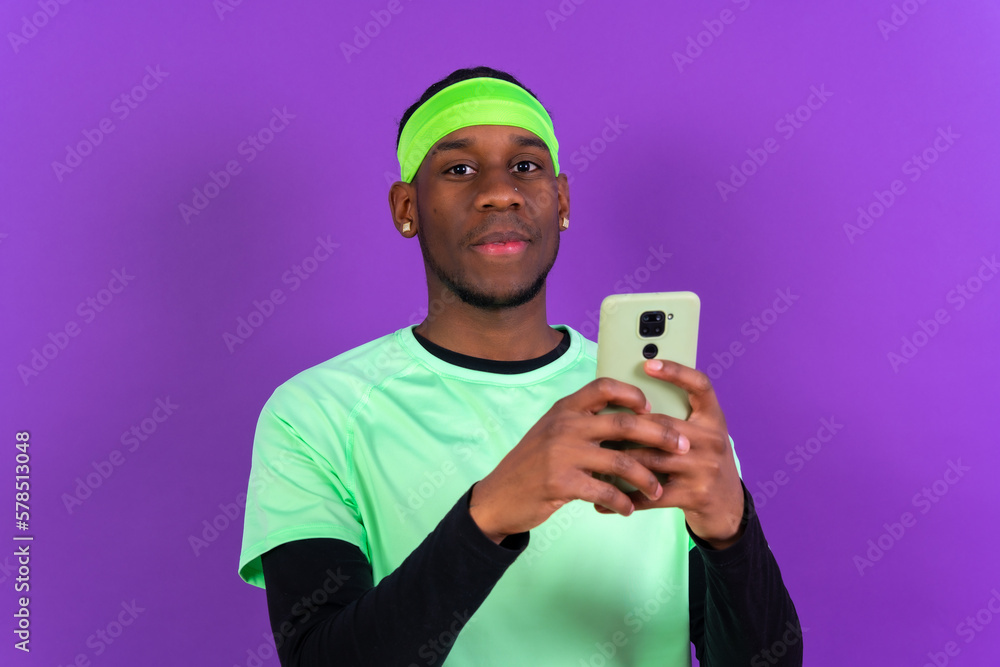 Black ethnic man holding a phone in green sportswear isolated on purple colored background Black ethnic man holding a phone in green sportswear isolated on purple colored background