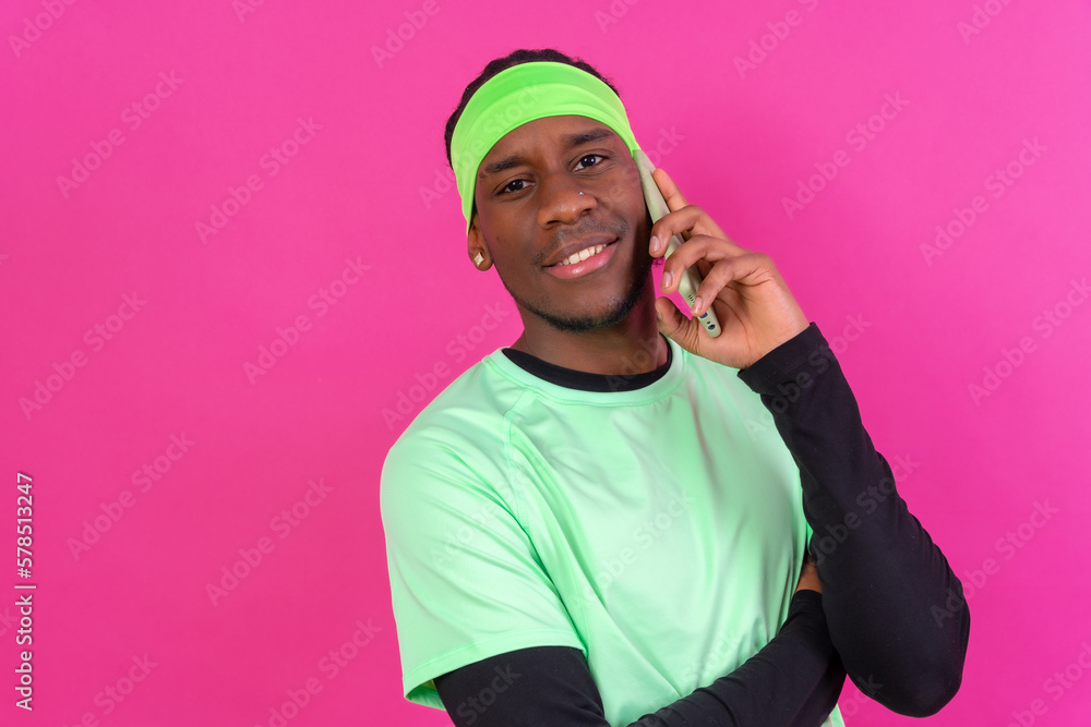 Fototapeta premium Black ethnic man with a phone in green clothes on a pink background, smiling talking on the phone