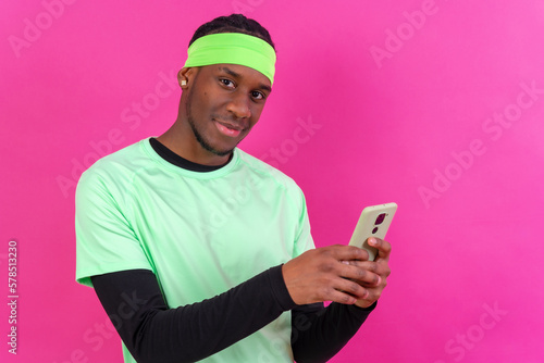 Black ethnic man with a phone in green clothes on a pink background, smiling