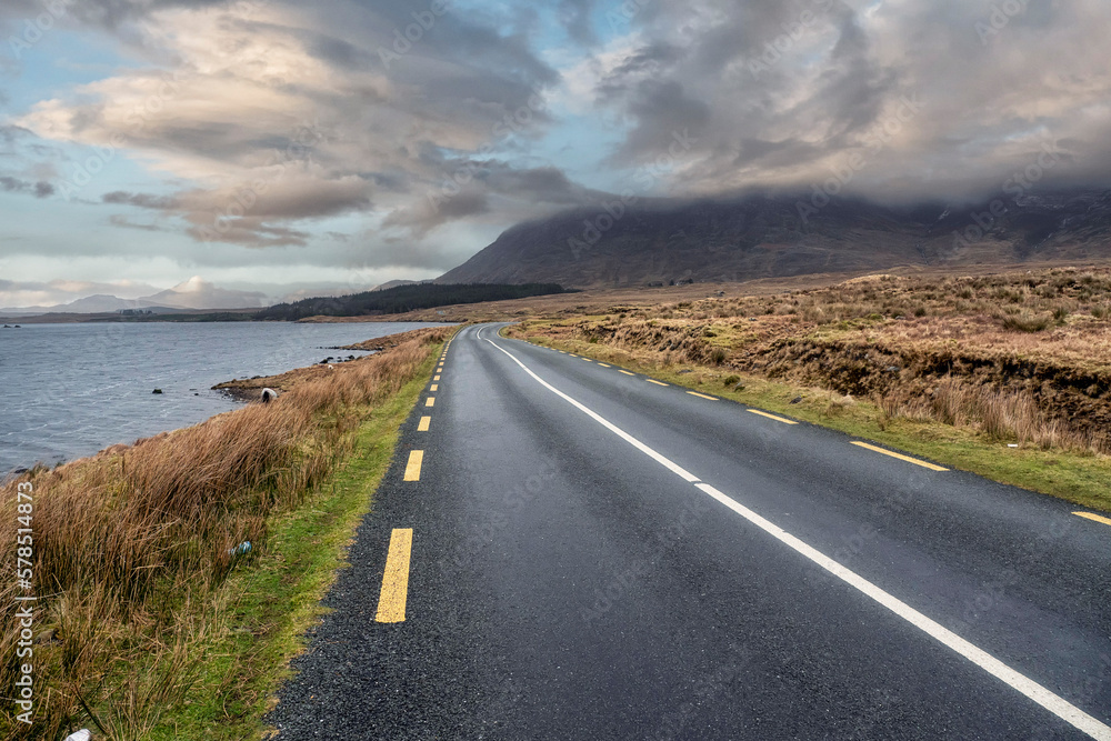 Beautiful scenery and small road by a lake in Connemara, Ireland. Mountains and cloudy sky in the background. Travel and transportation concept.