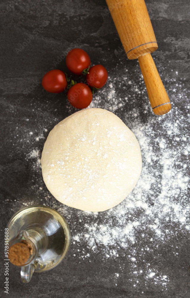 Background for making pizza, raw dough, cherry tomatoes, and a rolling pin for rolling out the dough on a dark background. Pizza cooking concept. Background for making pizza, raw dough, cherry tomatoes, and a rolling pin for rolling out the dough on a dark background. Pizza cooking concept.