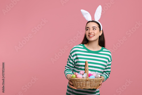 Happy woman in bunny ears headband holding wicker basket of painted Easter eggs on pink background. Space for text