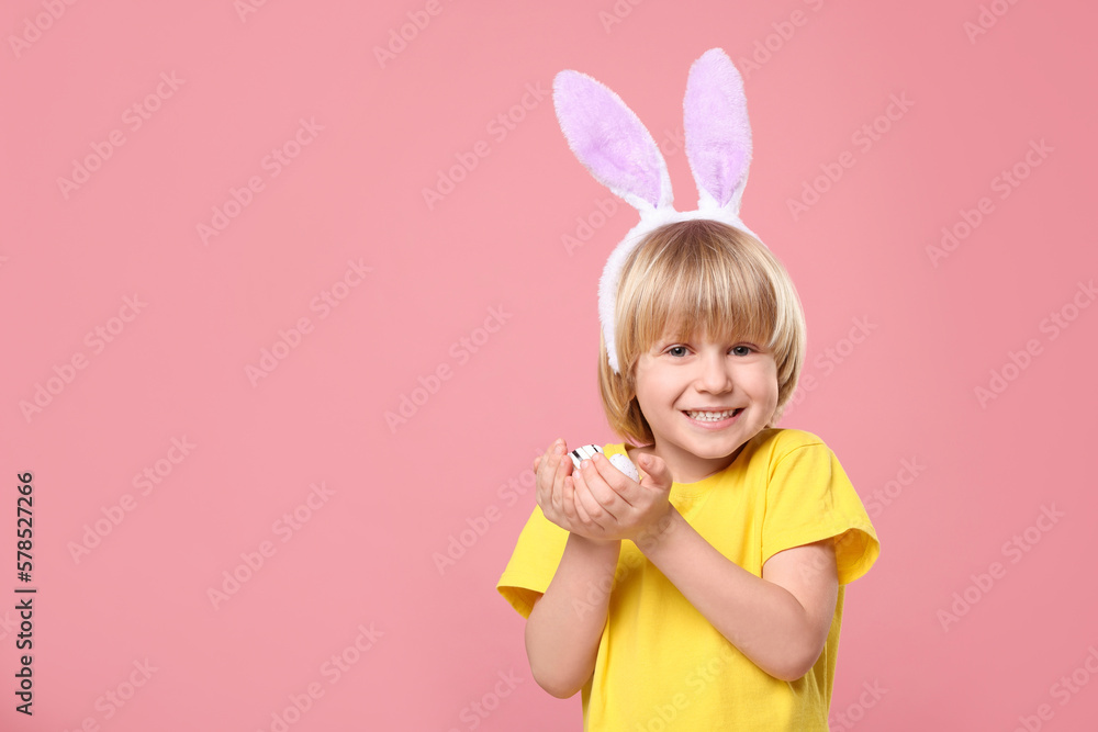 Happy boy in bunny ears headband holding painted Easter eggs on pink background. Space for text