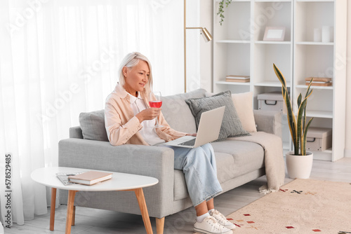 Mature woman with glass of wine using laptop on grey sofa at home