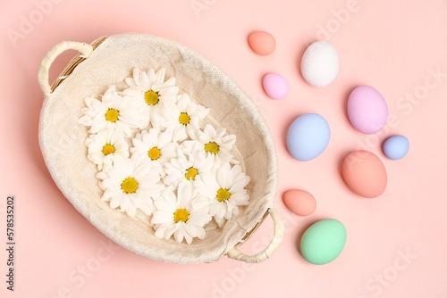 Wicker basket with chrysanthemum flowers and painted Easter eggs on pink background