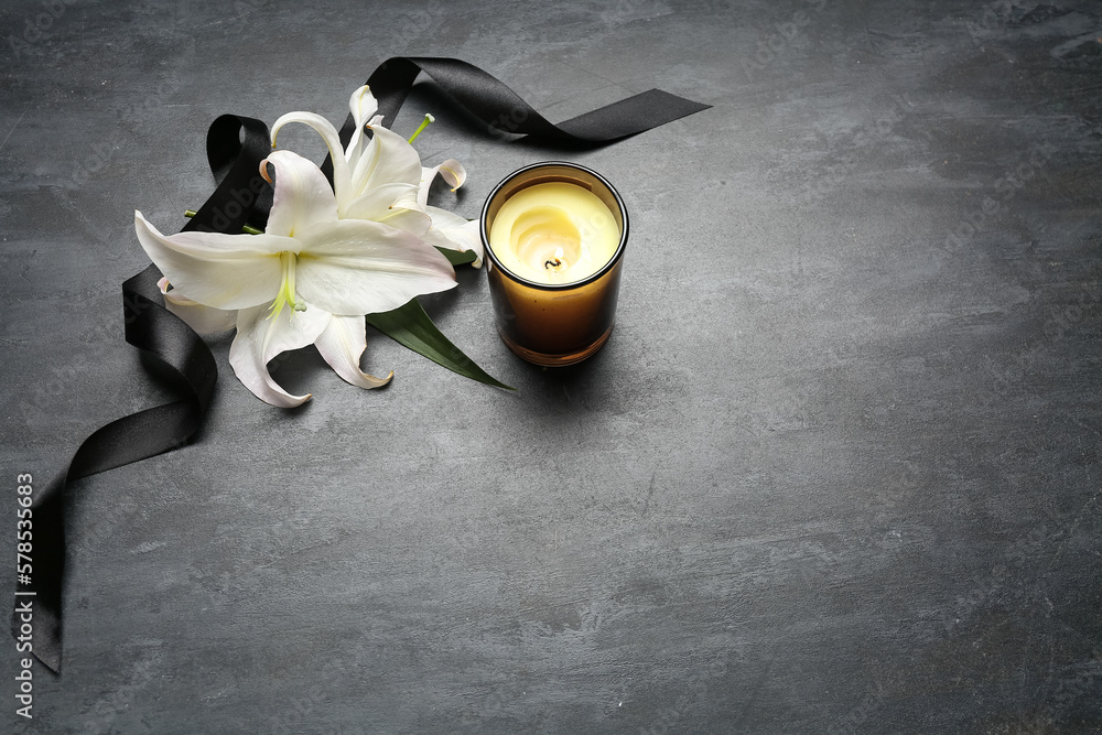 Composition with burning candle, white lily flowers and black funeral ribbon on dark background