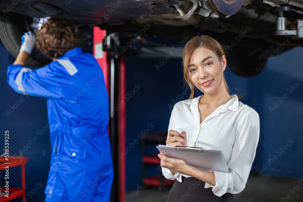 Portrait of young woman holding clipboard standing at workplace with mechanic in garage background. Female employees taking notes on repair work and accounting work of car service company.