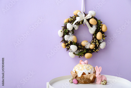 Table with Easter cake  rabbit and wreath on lilac wall