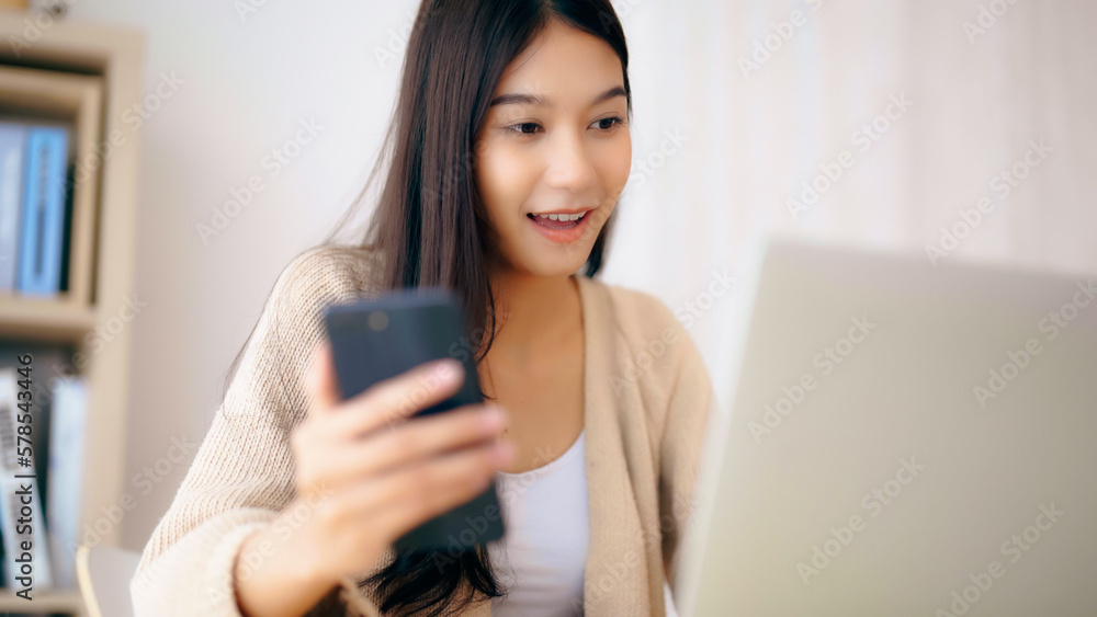 Young asian woman happy working on computer laptop and hand typing chat message on mobile smartphone at home. Video call, Online meeting Young asian woman happy working on computer laptop and hand typing chat message on mobile smartphone at home. Video call, Online meeting