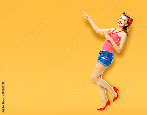Full body portrait of happy smiling beautiful woman. Pin up girl show, pointing finger, demonstrate, advertise. Vintage ad advertisement concept. Isolated over orange yellow background.