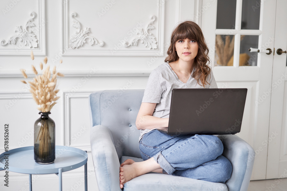 Fototapeta premium Woman Looks at the camera. Beautiful cheerful girl using black laptop while sitting on sofa in living room at home. Young casual woman using laptop for communication and work. Stay home