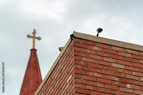 Brick wall with camero on edge watching pedestrians with background steeple and spire with golden cross on top