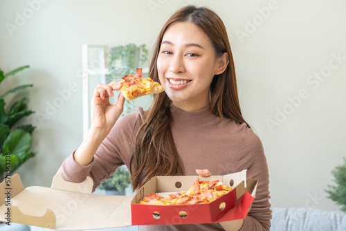 Happy meal with fast food, pleasure asian young woman, girl hold piece, enjoy eating delicious slice of pizza having takeaway at home relaxing resting, like taste good appetite, snack in lunch, dinner