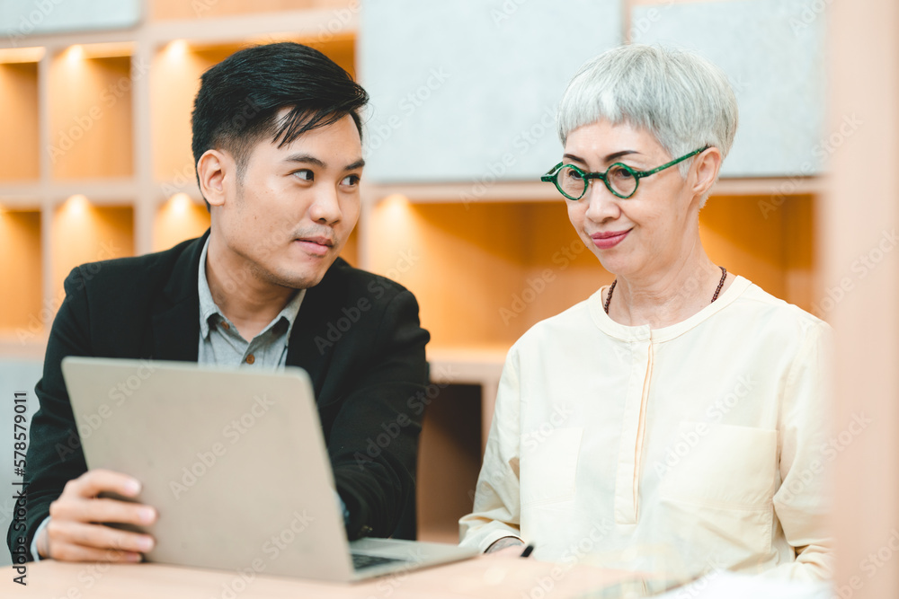 Fototapeta premium Young Asian business man in formal clothing showing something on digital tablet to senior female boss and manager wearing eyeglasses while in a meeting and training on technology