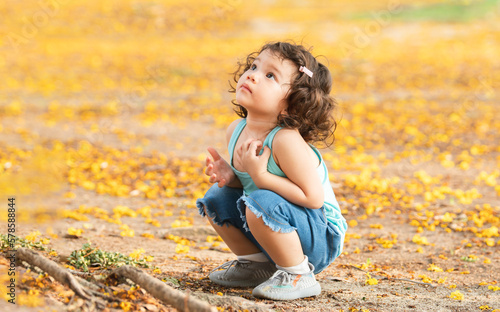 фотографія Little Caucasian child girl sitting under the tree, looking up to yellow leaves falling down on ground at park