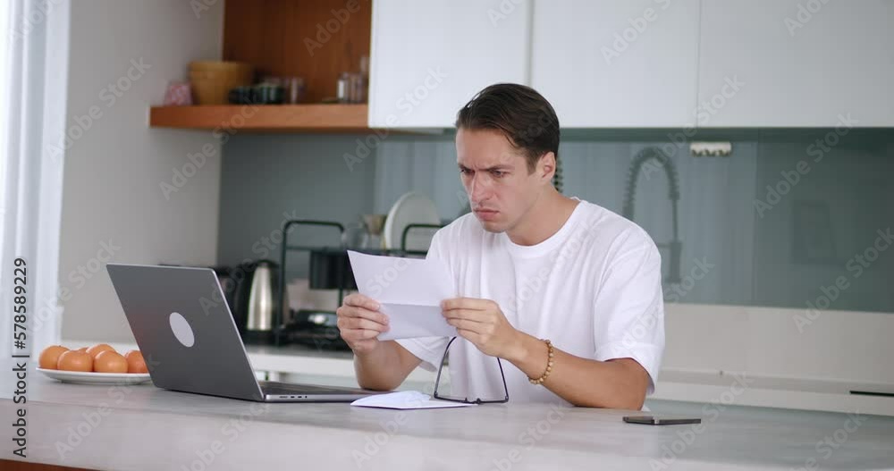 Frustrated man reading letter of rejection to accept job, sitting at table at home kitchen. Man frustrated by rejection tears up paper letter throws it on table, shows aggression. Concept of failure Frustrated man reading letter of rejection to accept job, sitting at table at home kitchen. Man frustrated by rejection tears up paper letter throws it on table, shows aggression. Concept of failure