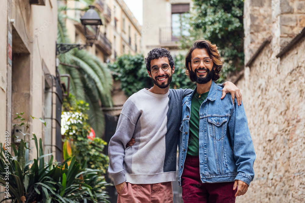 Naklejka premium gay male couple embracing looking at camera smiling happy in a nice street with plants, concept of leisure and love between people of the same sex