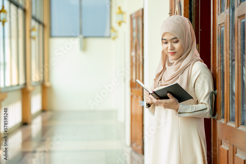 The image of an Asian Muslim woman in the Islamic religion in hijab in cream color. Standing reading the Qur'an and having a happy smiling face Staying in a beautiful mosque out of respect for God.