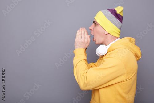 Side view of young adult man wearing yellow hoodie and beanie hat posing isolated over gray background  screaming with hands near mouth  making announce  yelling loud.