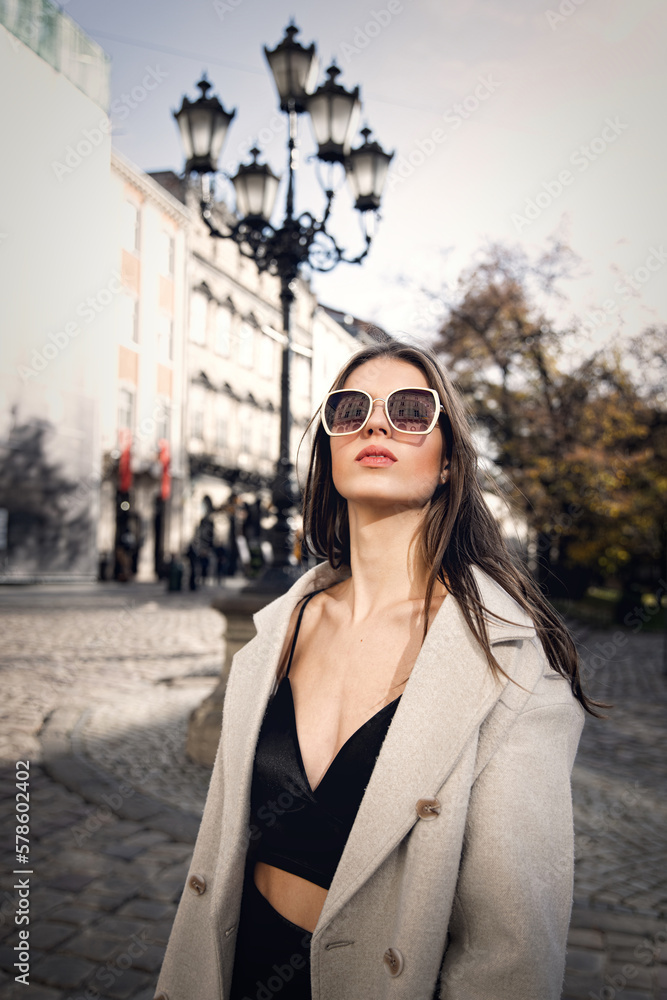 Fototapeta premium charming brunette posing on the street in spring, wearing a stylish beige coat, a top with razors and sunglasses
