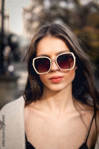 close-up portrait of a charming brunette on the street in spring, wearing a stylish beige coat, a top with razors and sunglasses © Mykola