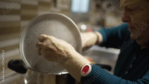 older woman in kitchen moving pot searing meat, video of grandmother talking and lifting lid of pot to turn food over, on her wrist she wears telecare bracelet