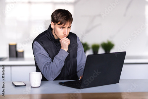 Handsome man using a laptop pc in the kitchen. Working from home in quarantine lockdown.