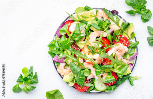 Delicious shrimp salad with avocado, cherry tomatoes, arugula, lamb lettuce, cucumber, red onion and sesame seeds on white table background. Top view