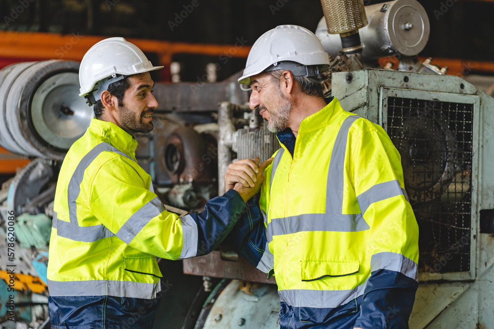Two senior male engineers and workers wearing safety vests and jacket along with hardhat or helmet while clenching hands and making a fist standing in front of machine in warehouse Two senior male engineers and workers wearing safety vests and jacket along with hardhat or helmet while clenching hands and making a fist standing in front of machine in warehouse