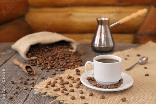 Against  the background of log wall  on a wooden table there is a white cup of coffee with a scattering of coffee beans from a small burlap bag  a copper turk and a wooden scoop with coffee beans. 