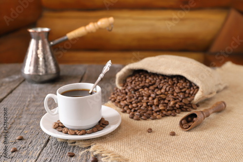 Against  the background of log wall  on a wooden table there is a white cup of coffee with a scattering of coffee beans from a small burlap bag  a copper turk and a wooden scoop with coffee beans. 