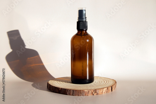 Layout of a brown glass bottle with an atomizer. The spray bottle stands on a round wooden podium on a beige background.
