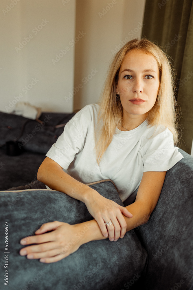 Serious blonde Caucasian girl sitting on sofa and looking curiously at camera Serious blonde Caucasian girl sitting on sofa and looking curiously at camera