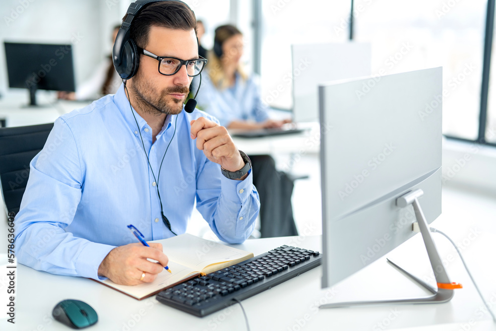 Serious successful business man wearing headset looking at computer monitor and taking notes to notebook accompanied with colleagues in the background. Serious successful business man wearing headset looking at computer monitor and taking notes to notebook accompanied with colleagues in the background.