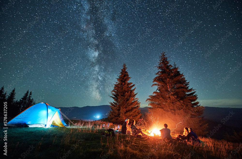 Night camping in mountains under starry sky. Group of people tourists having a rest near campsite, burning campfire and illuminated tent. Concept of tourism, hiking and adventure. - obrazy, fototapety, plakaty 