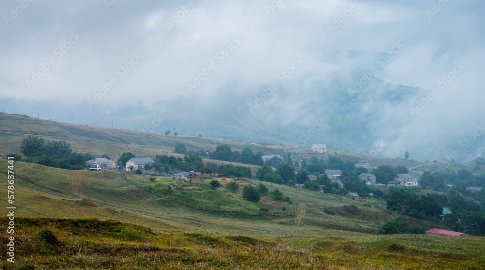 Landscape with a mountain village and fields with flowers on a cloudy summer day after rain with a haze.