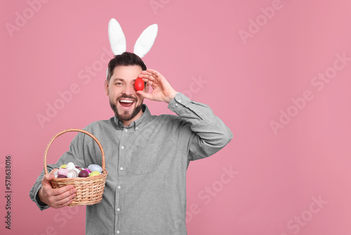 Happy man in bunny ears headband holding wicker basket with painted Easter eggs on pink background. Space for text