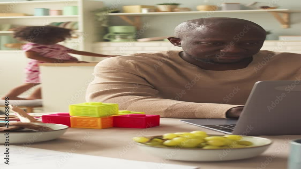 Vertical shot of African American businessman using laptop while working from home and his little daughter playing in background Vertical shot of African American businessman using laptop while working from home and his little daughter playing in background