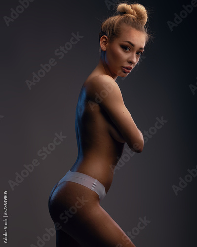 A beautiful half-naked woman covering chest with hands. Topless blonde model in white seamless panties covering breast by arms on a gray studio background.