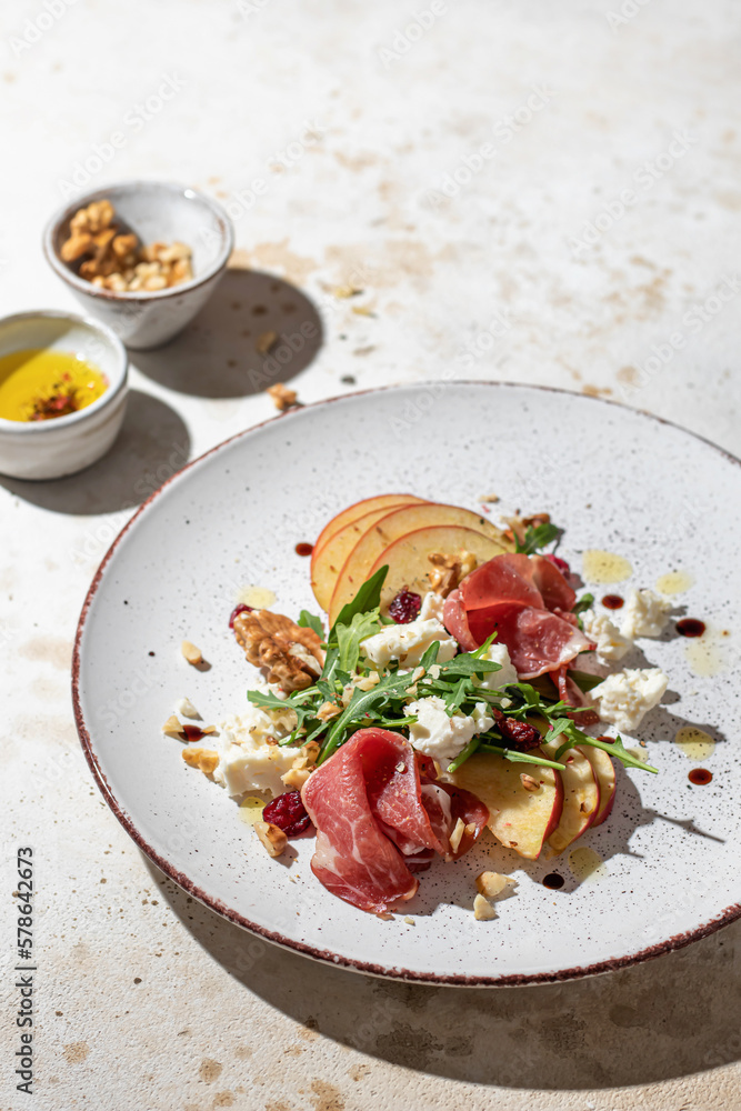 Salad of arugula and prosciutto with addition of sliced apple, cheese, walnuts and spices on a plate on light textured background in hursh light with clear shadows. Space for text