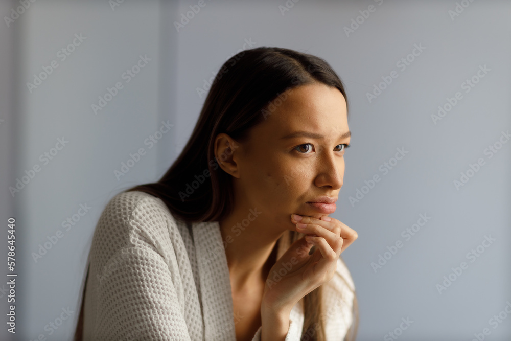 Attractive upset young woman in white bathrobe examines pimples on her face. Problematic skin on the face, acne. Portrait of girl removing pimples in the bathroom. Beauty and health of the skin. Attractive upset young woman in white bathrobe examines pimples on her face. Problematic skin on the face, acne. Portrait of girl removing pimples in the bathroom. Beauty and health of the skin.