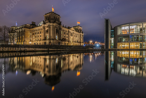 View of government buildings at night time. Reichstag in Berlin at the blue hour. Illuminated historical buildings. River Spree with reflection on water surface in winter