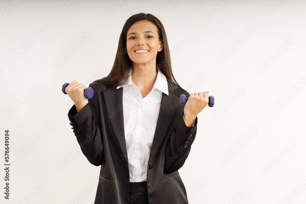 Attractive girl with dark hair in stylish business suit and white shirt holds small female dumbbells. Portrait of happy smiling young woman with sports equipment on isolated white background. Attractive girl with dark hair in stylish business suit and white shirt holds small female dumbbells. Portrait of happy smiling young woman with sports equipment on isolated white background.
