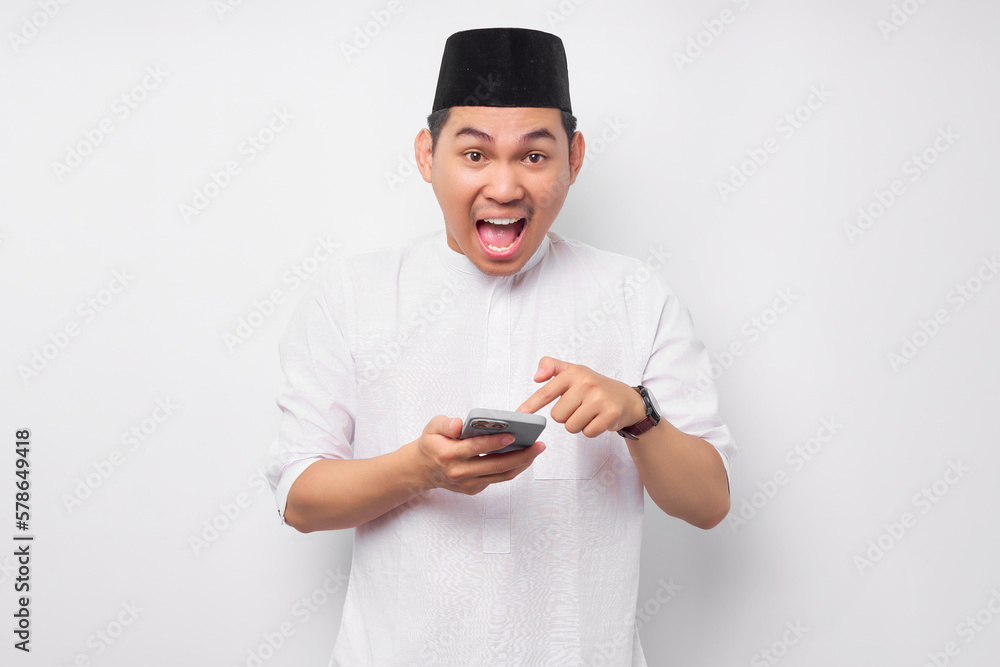 Excited young Asian Muslim man wearing Arabic clothes pointing finger at smartphone isolated on white background. People religious Islamic lifestyle concept Excited young Asian Muslim man wearing Arabic clothes pointing finger at smartphone isolated on white background. People religious Islamic lifestyle concept