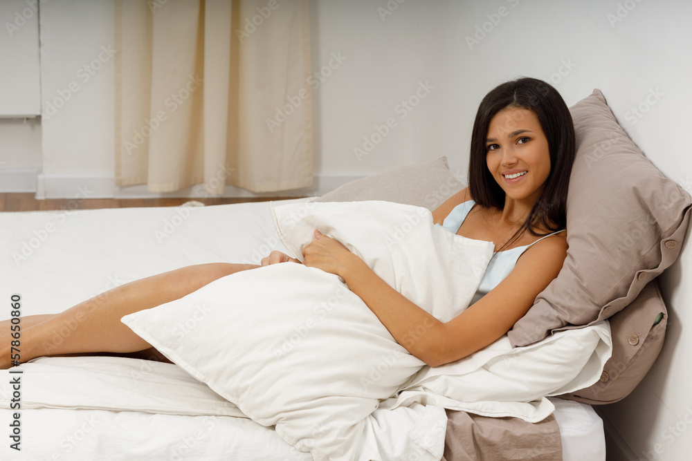 Beautiful slender young woman, dressed in gently light blue silk nightie negligee, lies in cozy bed in the bedroom. Attractive girl hug pillow after waking up from sleep. Good morning. Beautiful slender young woman, dressed in gently light blue silk nightie negligee, lies in cozy bed in the bedroom. Attractive girl hug pillow after waking up from sleep. Good morning.