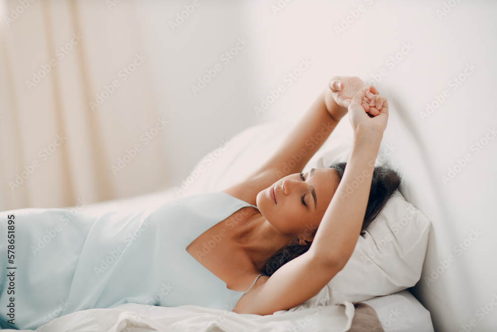 Fototapeta premium Beautiful slender young woman, dressed in gently light blue silk nightie negligee, lies in cozy bed in the bedroom. Attractive girl stretches after waking up from sleep. Good morning.