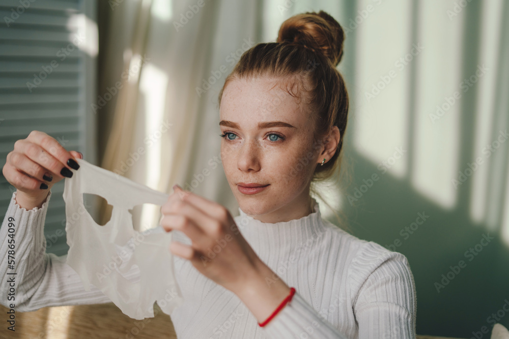 Beautiful young woman with red hair and freckled face applying facial sheet mask on her face, having self care day at home. Wellbeing concept. Skin care and Beautiful young woman with red hair and freckled face applying facial sheet mask on her face, having self care day at home. Wellbeing concept. Skin care and