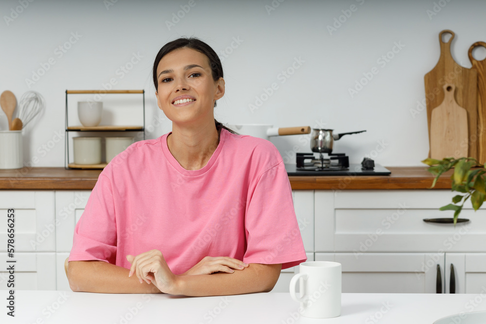 Cute young brunette woman in casual clothes looks straight into the camera. Portrait of smiling happy girl sitting at home in the kitchen at the dining table Cute young brunette woman in casual clothes looks straight into the camera. Portrait of smiling happy girl sitting at home in the kitchen at the dining table